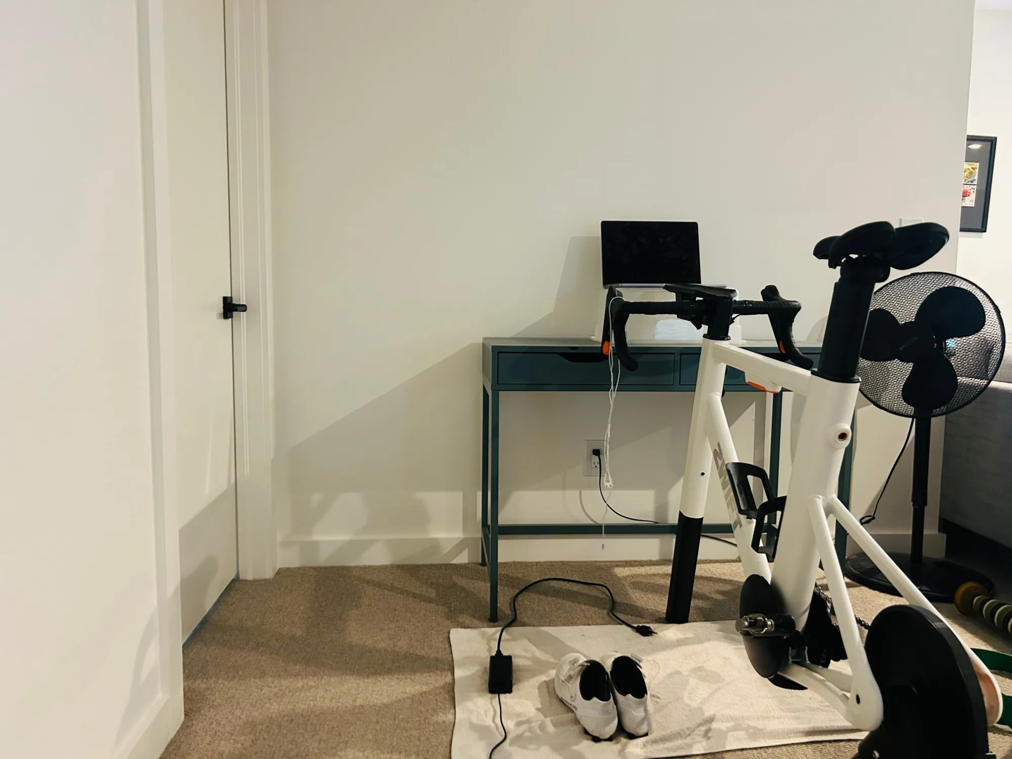 A pain cave ready to go: a bike on a smart trainer, a fan, a laptop on a plain desk, a towel on the floor and cycling shoes waiting
