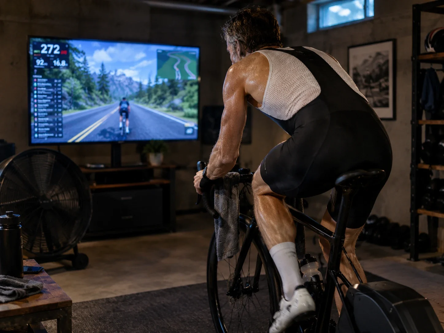 A sweaty rider mid effort on an indoor trainer in a basement, eyes on Zwift, with a fan and a towel on the bars