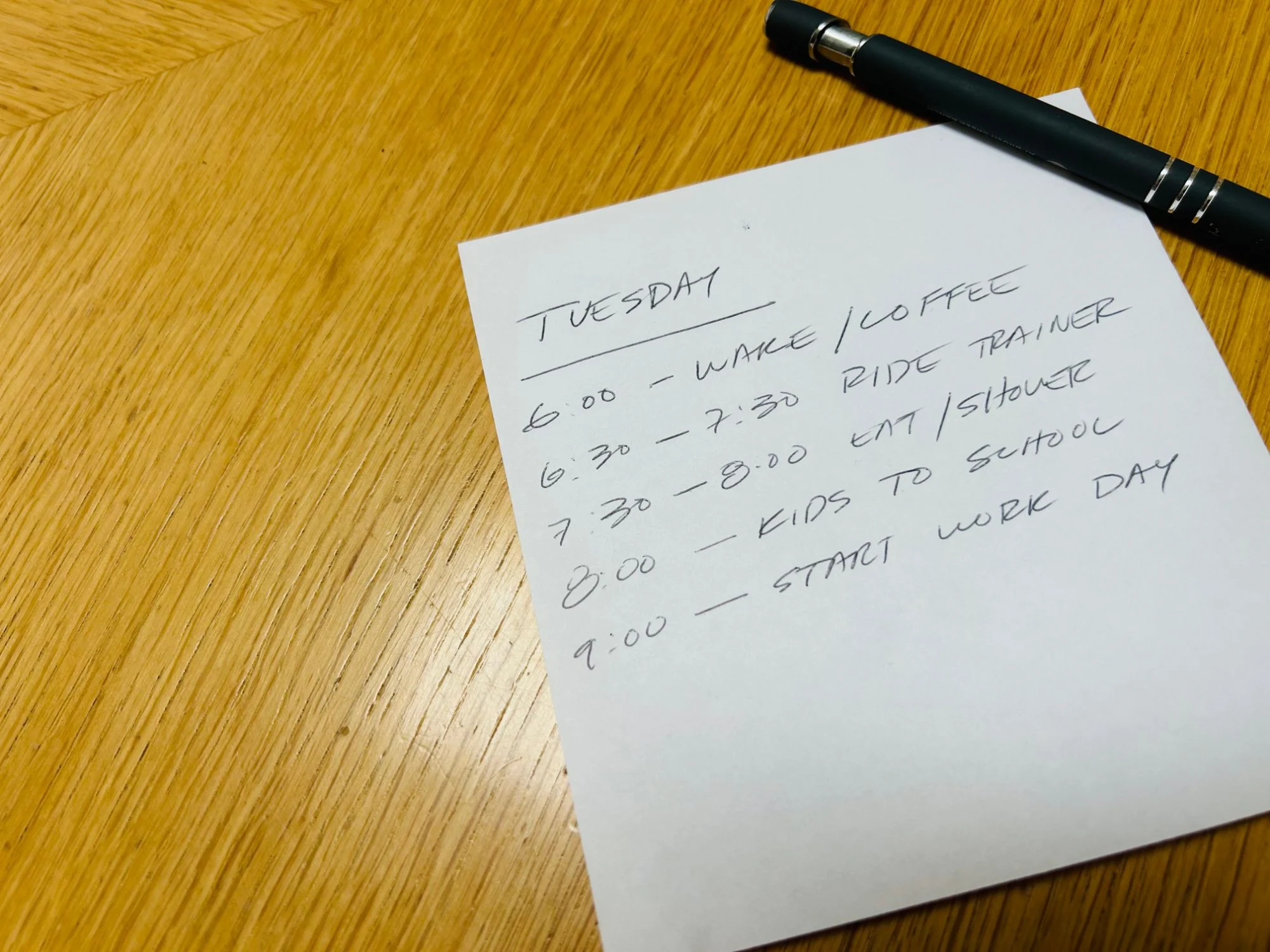 A handwritten Tuesday plan on a wooden table: 6:00 wake and coffee, 6:30 to 7:30 ride trainer, 7:30 to 8:00 eat and shower, 8:00 kids to school, 9:00 start work day